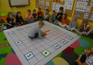 Grupa dzieci siedzi wokół rozłożonej na dywanie macie do kodowania. Na macie ułożony jest zegar z tabliczek oraz z papierowych wskazówek. Chłopiec przestawia na zegarze wskazówki.